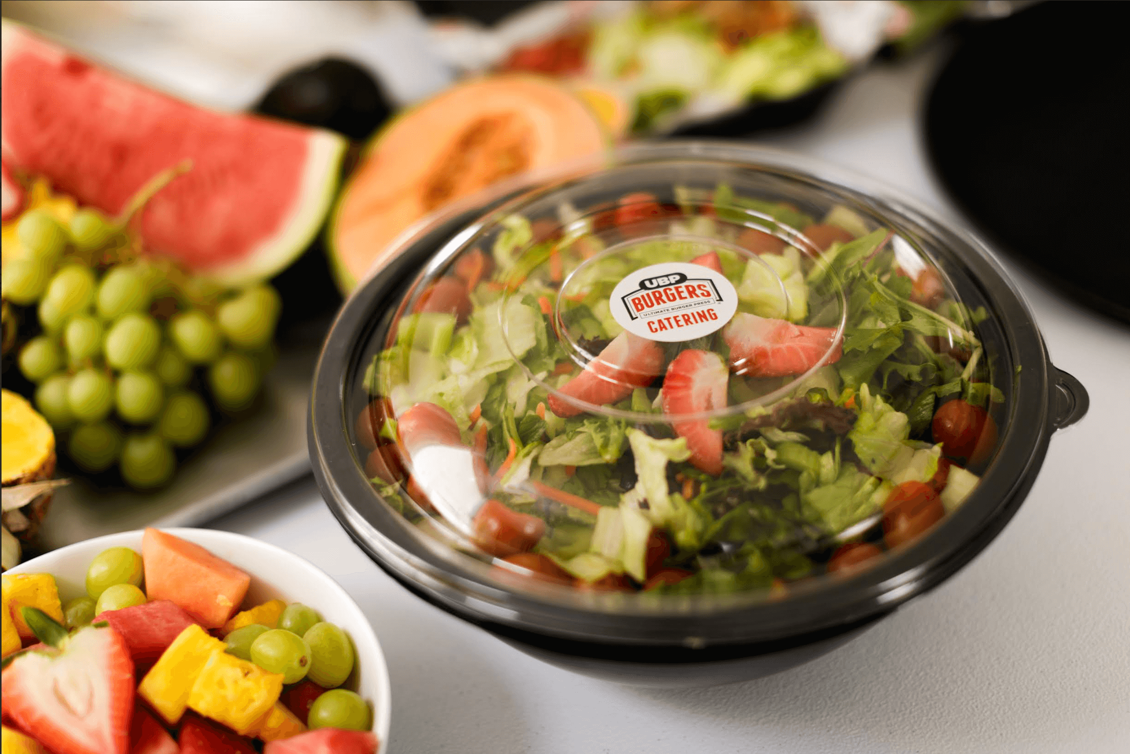 UBP Burgers salad in a clear container on a table with fruits and vegetables.